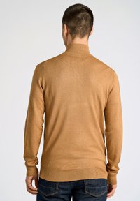 Maglione lungo beige a maniche lunghe con colletto a coste, design aderente e texture liscia, abbinato a jeans di denim scuro e dettagli visibili sul retro.