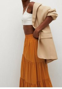 Blazer beige indossato sopra un top bianco corto e una gonna lunga arancione a balze. Il tessuto appare morbido con una texture fluente e dettagli arricciati.