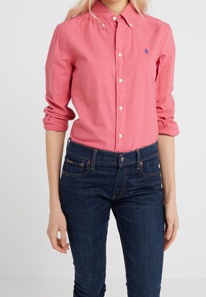 Kvinde iført en koralpink skjorte med knapper og op-rullede ærmer samt mørkeblå jeans foran en ensfarvet baggrund.
