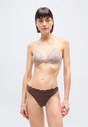 Femme aux cheveux courts et foncés portant un bralette en dentelle beige et une culotte marron foncé bordée de dentelle, debout devant un fond clair uni.