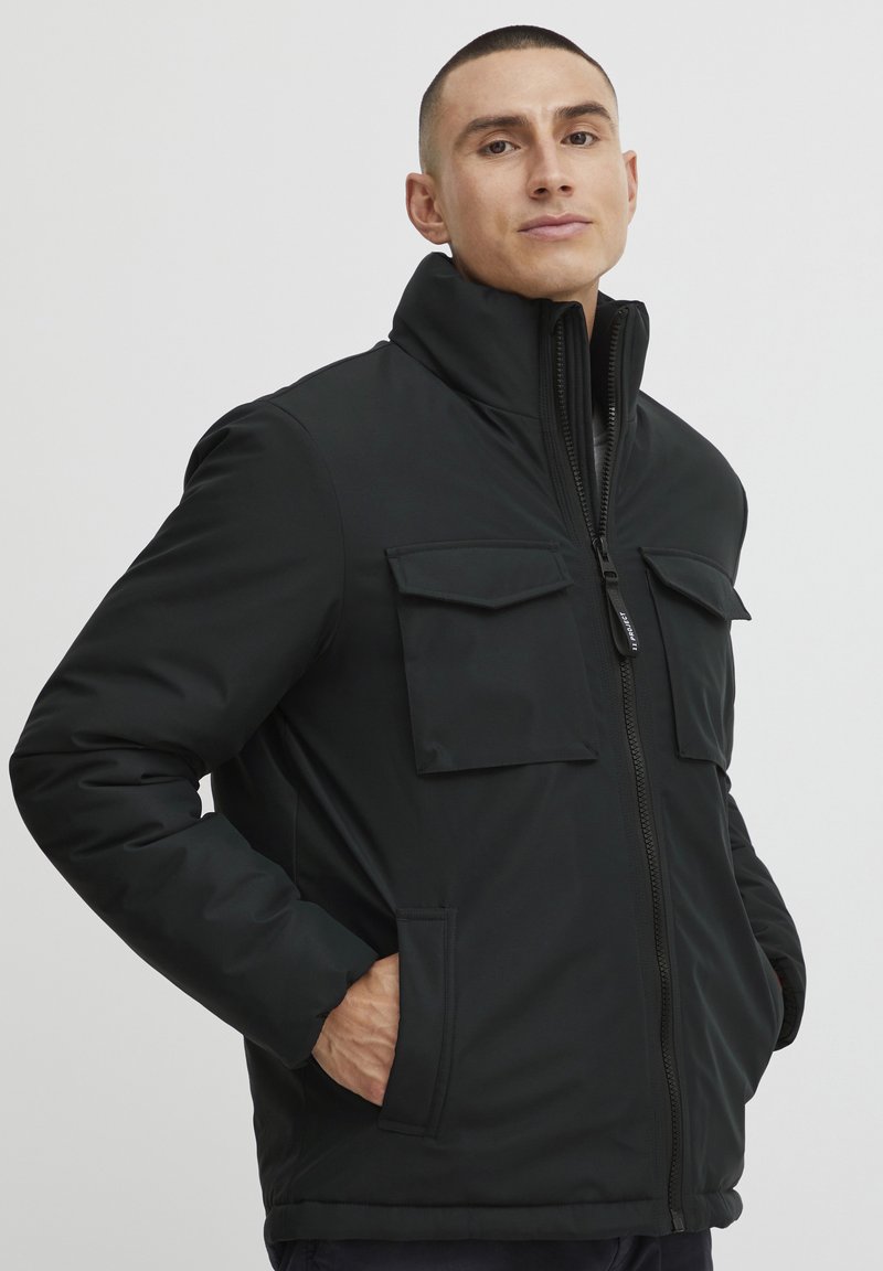 Chaqueta aislante negra con cuello alto, cierre frontal de cremallera y dos bolsillos en el pecho. El material parece suave con un acabado mate.