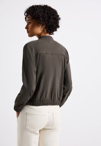 Olivgrön bomberjacka med en samlad fåll, långa ärmar och en slät textur. Har en central söm bak och diskret märkning.