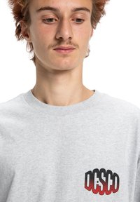 Šedé bavlněné tričko s kulatým výstřihem a krátkými rukávy. Černé a červené grafické logo v oblasti levého prsa. Standardní střih.