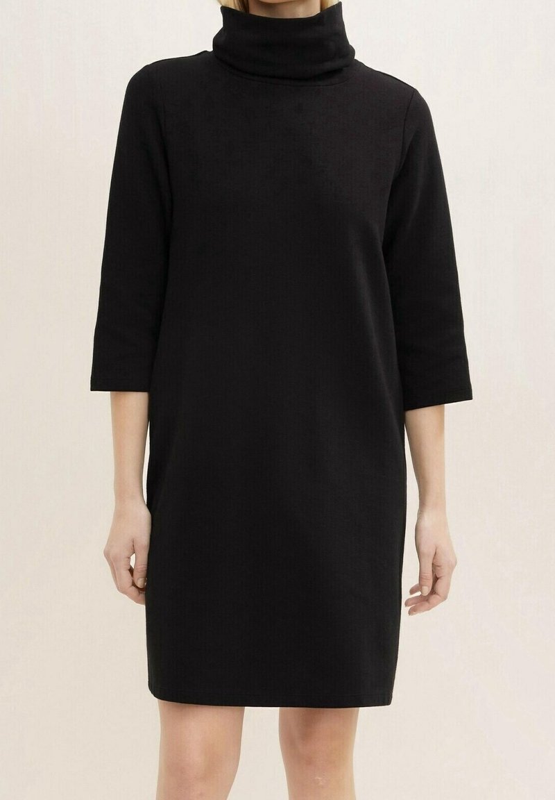 Femme vêtue d'une robe noire simple au genou avec un col bénitier haut et des manches trois-quarts, debout devant un fond clair uni.