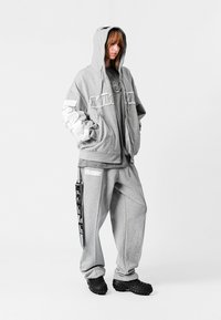 Sudadera gris de gran tamaño con acentos blancos, combinada con pantalones de chándal grises que tienen bolsillos laterales y una etiqueta. Calzado negro completa el atuendo.