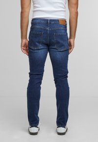 Jeans en denim bleu foncé avec une coupe slim, design à cinq poches, et un patch en cuir marron sur la ceinture arrière. Lavable en machine.