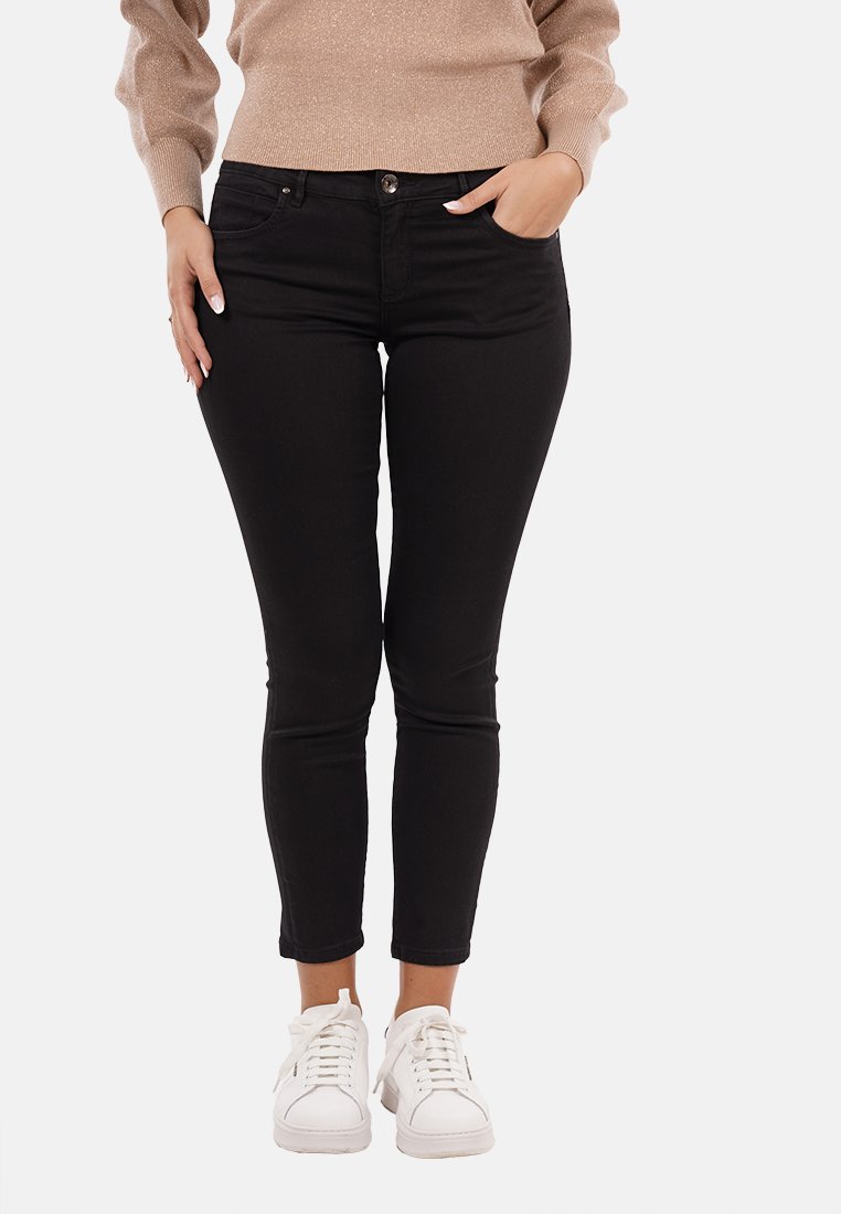 Donna che indossa jeans skinny neri, sneaker bianche e un top beige a maniche lunghe, in piedi con una mano in tasca e l'altra lungo il corpo.