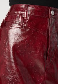 Bordeauxfarbene Kunstlederrock mit glänzender Oberfläche, ausgestattet mit einem Fünf-Taschen-Design, Reißverschluss und sichtbaren Nähten.
