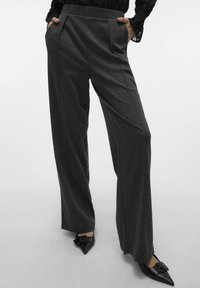 Pantaloni a gamba larga in tessuto grigio scuro, con una superficie testurizzata, pieghe frontali e tasche laterali, abbinati a scarpe nere a punta.