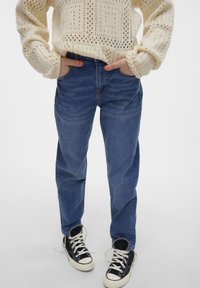 Ljusgrå stickad tröja med geometriska hål, matchad med medelmörka blå raka jeans och svarta hög-top sneakers med vita sulor.