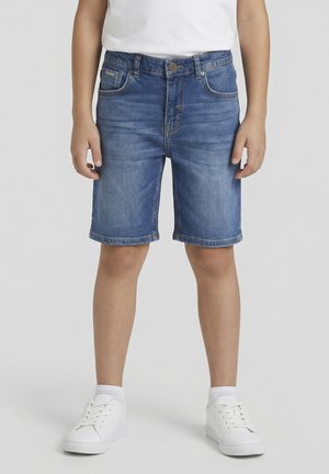 Niño con pantalones cortos de mezclilla azules hasta la mitad del muslo, zapatillas blancas y camisa blanca, de pie con las manos relajadas a los lados.