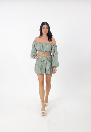 Kobieta o długich ciemnych włosach, ubrana w zielony kwiatowy krótki top z odkrytymi ramionami i pasujące szorty, stoi na białym tle.