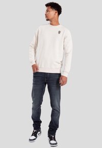 Sudadera beige claro con cuello redondo, pequeño gráfico negro en el pecho, combinada con jeans azul oscuro y zapatillas en blanco y negro.
