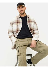 Geruite flanellen shirt in crème, rood en bruine tinten, gelegd over een zwart T-shirt, met beige cargobroek met ritszakken.