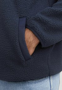 Marineblå fleece-pullover med tekstureret overflade; har en kontrasterende stoflomme; afslappet pasform med en synlig hånd, der hviler inden i lommen.