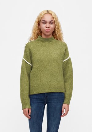 Jeune femme aux longs cheveux blonds bouclés portant un pull en maille vert et un jean bleu, debout devant un fond blanc uni.