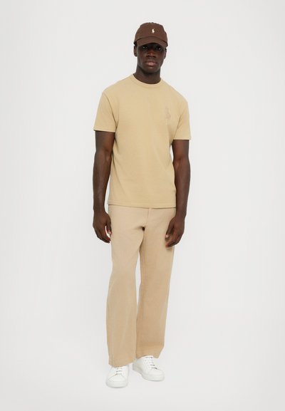 Beige bomullströja med liten logotyp, i kombination med beiga vida byxor och vita sneakers. En brun keps avslutar outfiten.