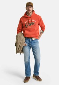 Rode hoodie met "Gant" logo, beige jas over de arm, lichtblauwe spijkerbroek, bruine schoenen. Hoodie heeft een kangoeroezak en trekkoorden.