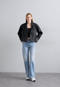 Schwarze Leder-Bikerjacke, tailliert mit breiten Revers, kombiniert mit hellblauen Schlagjeans und weißen Sneakers, vor einem grauen Hintergrund.