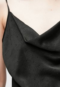 Zwart top met kraag en textuur van kleine zwarte versieringen. Dunne bandjes en een zachte val benadrukken het silhouet.