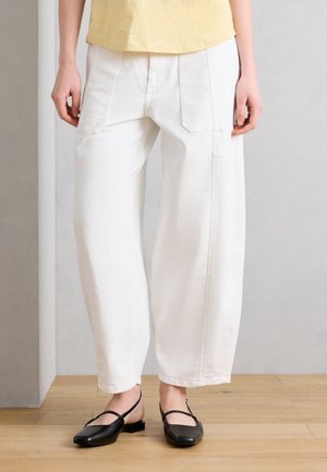 Személy laza, fehér nadrágot visel nagy első zsebekkel és fekete, alacsony sarkú Mary Jane cipőkkel, egy fa padlón állva.