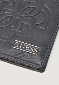 Czarna skórzana portmonetka z wytłoczonym logo, srebrną metalową metką "GUESS" oraz kontrastowym przeszyciem dla dodatkowego wykończenia.