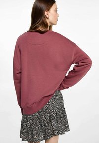 Felpa oversize color bordeaux con scollatura rotonda, texture morbida e spalle scese, abbinata a una gonna a fantasia nera e crema.