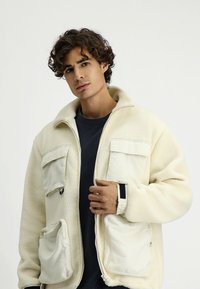 Jasno beżowa polarowa kurtka z dwiema przednimi kieszeniami naszytymi i jedną kieszenią na zamek błyskawiczny. Posiada wysoki kołnierz i ściągacze przy mankietach dla lepszej izolacji.