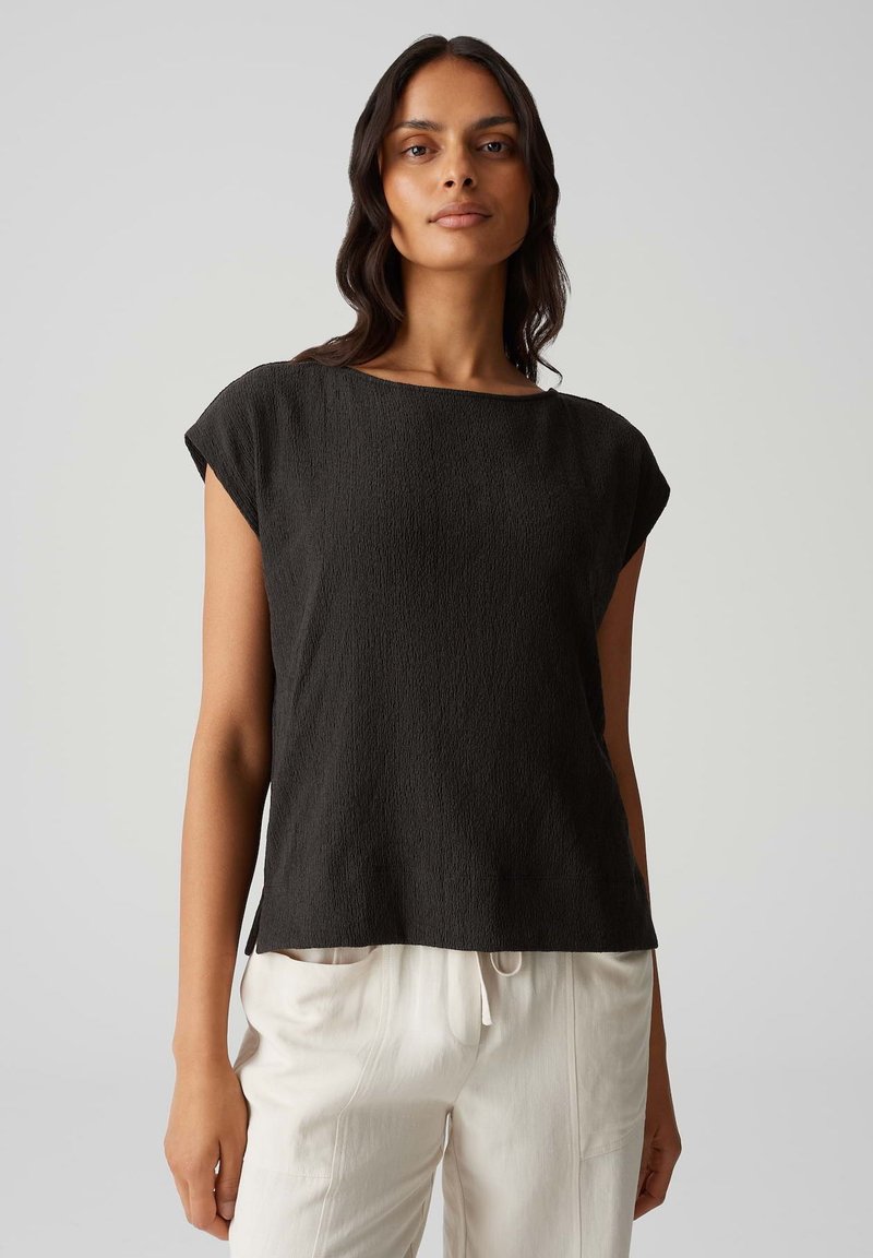 Zwarte, gestructureerde blouse met korte mouwen en een brede halslijn, gecombineerd met lichte, ruime broeken. Eenvoudig, minimalistisch ontwerp.