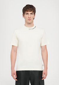 Hvit bomulls t-skjorte med rund hals, korte ermer og "EA7" logo og tekst brodert på øverste venstre side. Enkel, minimalistisk design.