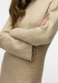 Pull en tricot côtelé beige à manches longues, avec un col haut et une texture douce. Le tissu présente des variations tonales subtiles.