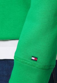 Grön sweatshirttyg med ribbstickade manschetter, som har en liten logopatch i rött, vitt och blått på ärmen. Slät textur, avslappnad design.