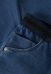 Pantalones de mezclilla en azul oscuro con una cinturilla elástica negra. Presenta un pequeño acento metálico en forma de corazón en el bolsillo y una tela texturizada.