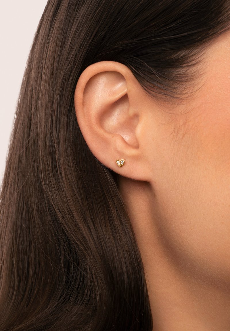 Orecchino a perno a forma di cuore in oro con piccoli gemme trasparenti, in primo piano su un orecchio e capelli lisci e scuri.