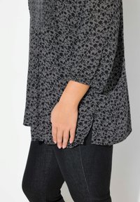 Haut gris à motifs floraux avec une texture smockée, manches trois-quarts et fentes sur les côtés, associé à un jean sombre.