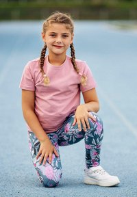 Różowa koszulka z krótkim rękawem w połączeniu z niebieskimi legginsami w kwiatowy wzór. Dziecko klęczy na nawierzchni toru, ubrane w białe sportowe buty.