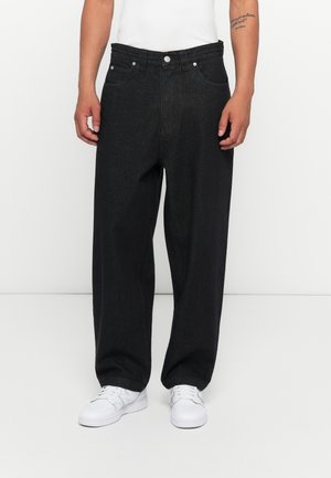 Mężczyzna w luźnych czarnych dżinsach z pięcioma kieszeniami i białych tenisówkach, stojący na tle jednolitego białego tła.