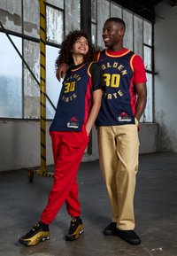 Due individui in abbigliamento sportivo: uno indossa una maglia blu navy con dettagli dorati e pantaloni rossi, l'altro indossa una maglia rossa e dorata con pantaloni beige.