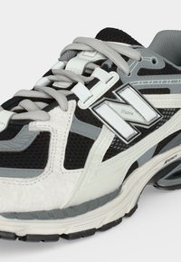 Κοντινή λήψη ενός αθλητικού παπουτσιού σε μαύρο, λευκό και γκρι με κορδόνια, από ύφασμα mesh και δερμάτινο υλικό, με μεγάλο λογότυπο "N" στο πλάι.