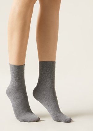 Piernas con calcetines tobilleros grises lisos, un pie apoyado completamente en el suelo y el otro ligeramente levantado sobre las puntas de los dedos, sobre un fondo neutro.