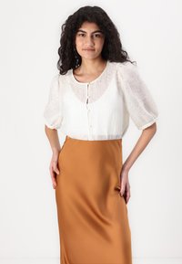 Blusa bianca testurizzata con maniche a sbuffo, scollatura rotonda e dettagli con bottoni, abbinata a una gonna lunga in satin marrone liscia.