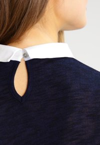 Top smanicato blu navy con colletto bianco, caratterizzato da un'apertura a chiave sul retro e chiusura con bottone. Texture liscia con una leggera finitura mélange.