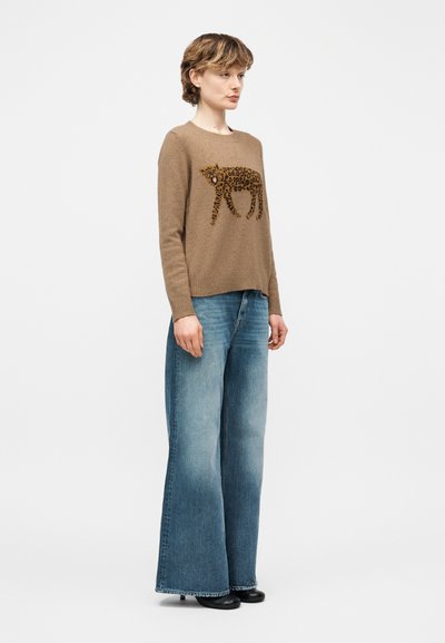 Femme debout en jean à jambes larges bleu et un pull marron avec un grand motif léopard sur le devant, portant des chaussures noires.