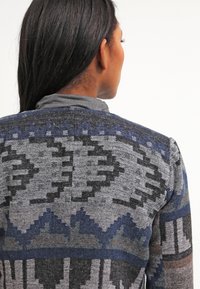 Femme aux longs cheveux noirs portant un pull en tricot à motifs gris et bleus, vue de dos sur un fond blanc.