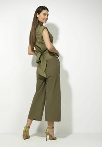 Olijfgroene jumpsuit met korte mouwen, strikdetail bij de taille en wijde pijpen. Gecombineerd met strappy groene hakken. Schone achtergrond.