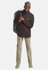 Veste softshell marron, pantalon khaki clair et chaussures de randonnée grises. Sac à dos avec un motif noir et gris. Sujet souriant tenant les bretelles du sac à dos.
