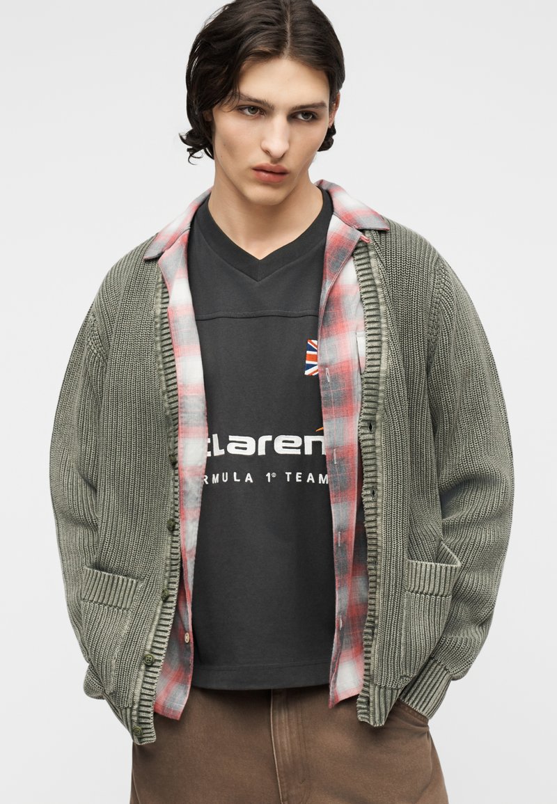 Junger Mann, der eine grüne Strickjacke über einem roten karierten Hemd und einem schwarzen McLaren Formel-1-T-Shirt trägt, braune Hose, Hände in den Taschen.