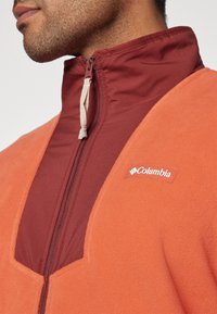 Giacca in pile con colletto alto, di colore arancione e bordeaux, con chiusura a zip, tessuto testurizzato e logo Columbia sul petto.