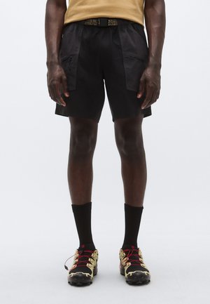 Mustad lühikesed püksid, kerged ja tekstuursed, millel on tõmblukuga taskud ja mustriline vöö. Kandmisega koos mustad sokid ja kollaste detailidega jalanõud.