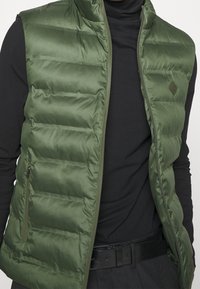 Pufferväst i djupgrön, med dragkedja, vadderad design och en sidficka. Bärs över en svart turtleneck för kontrast.